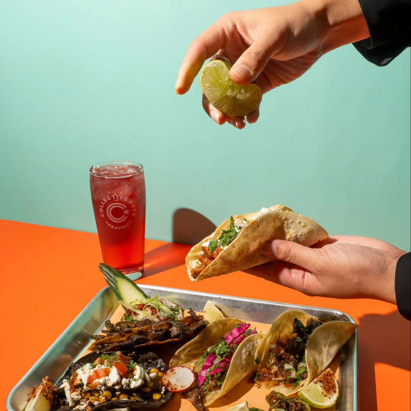 Person squeezing a fresh lime over a tray of authentic Mexican tacos with a cold beer in the background